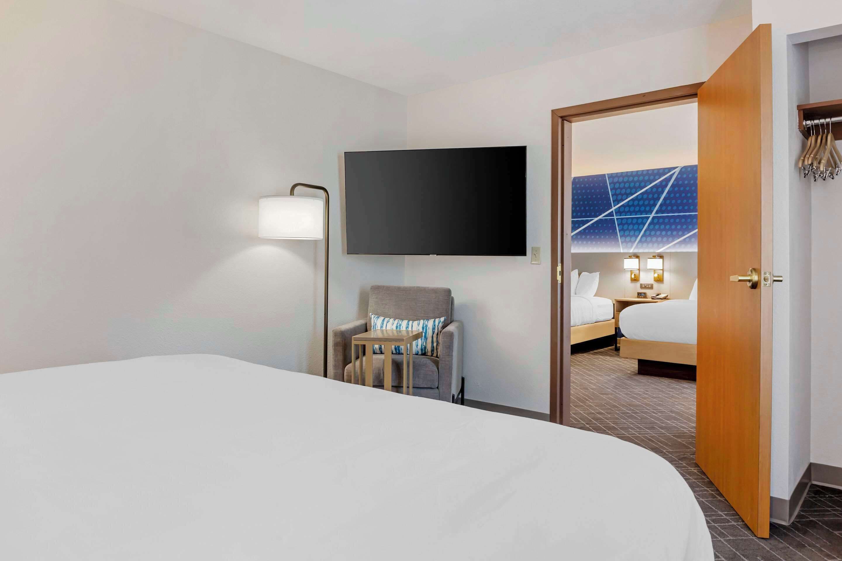 A hotel room with a large bed, a chair with a table, a flat-screen TV, and a door leading to another room with a bed and a bathroom.