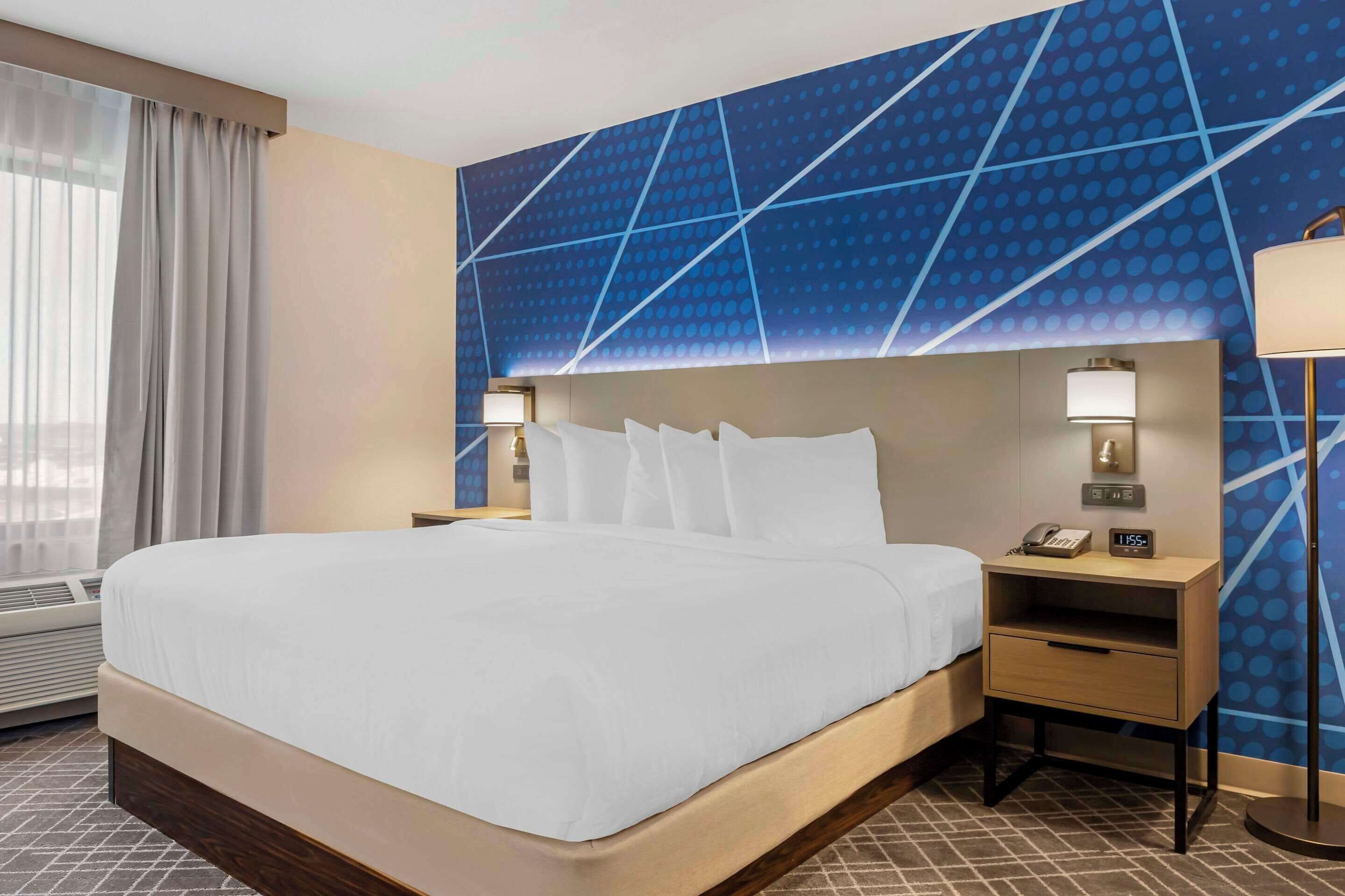 Una habitación de hotel con una cama grande, una mesita de noche con lámpara, un teléfono y un reloj digital.