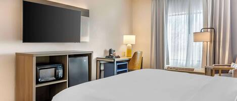 Habitación de hotel con una cama grande, un televisor de pantalla plana montado en la pared, un escritorio con lámpara, un microondas y una silla.