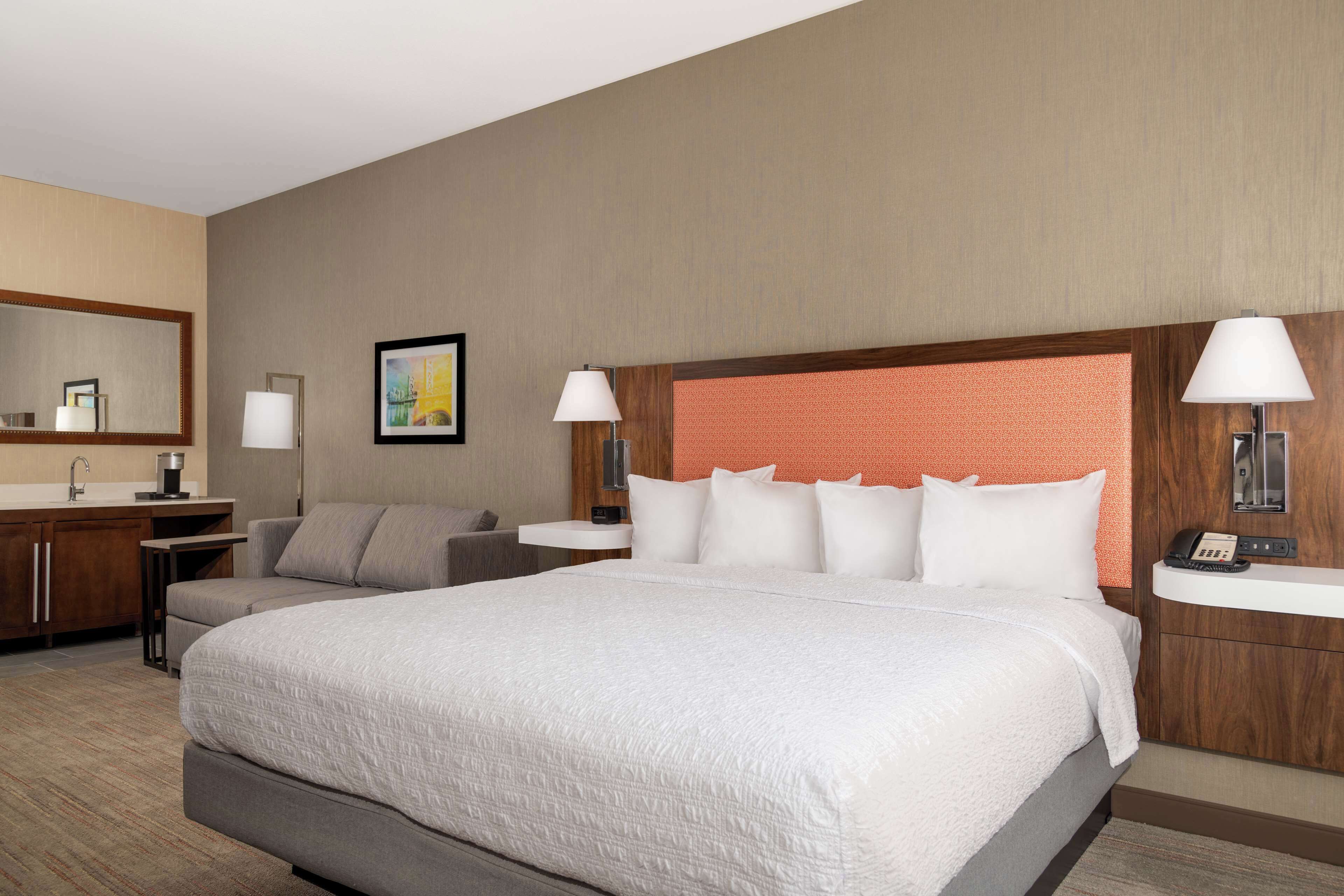 A hotel room with a large bed, two bedside tables, a nightstand, a sofa, a desk, a mirror, and a picture on the wall.