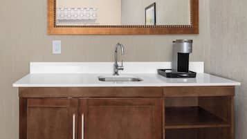 A modern bathroom with a white countertop, a stainless steel sink, a coffee maker, and a wooden cabinet.