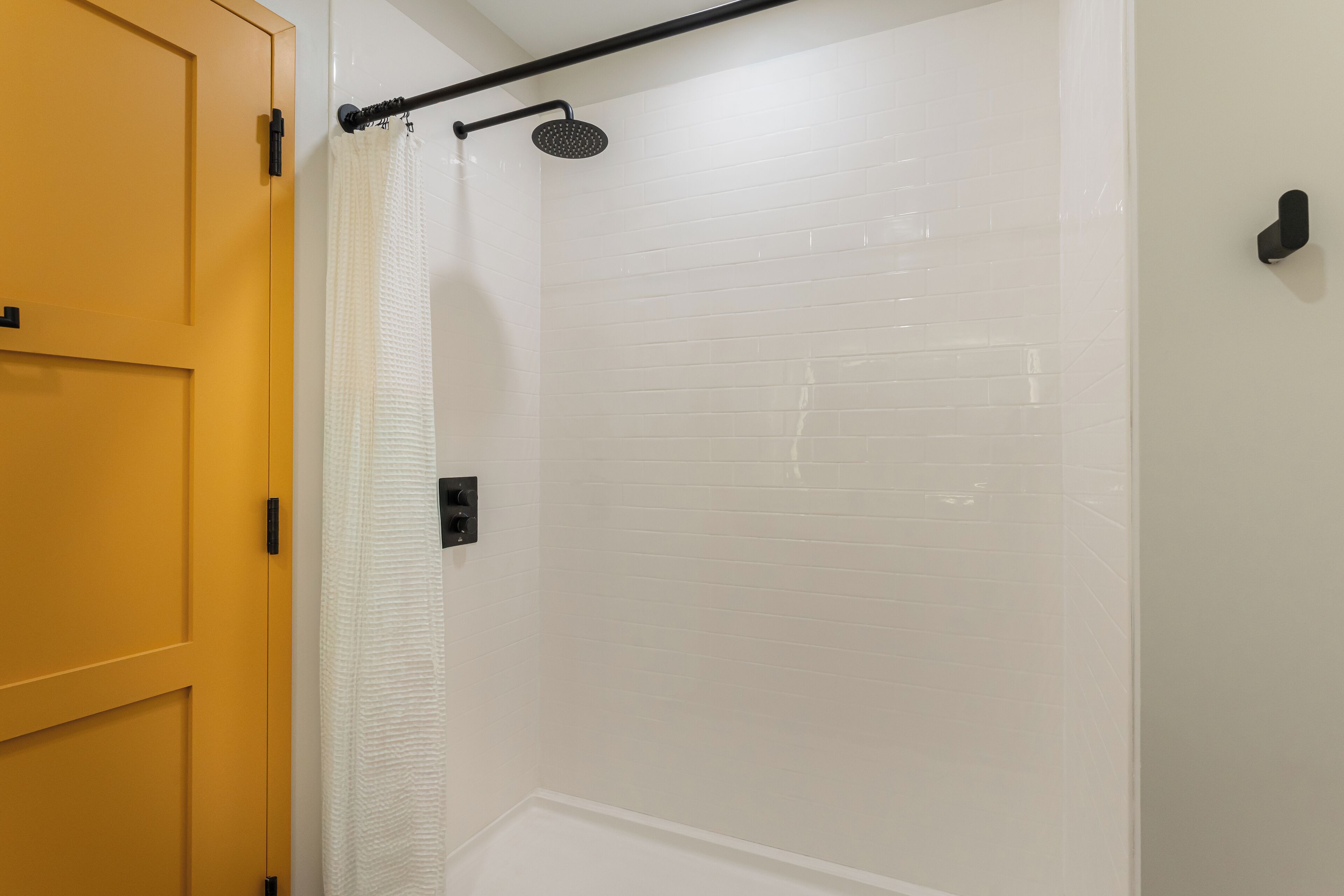 Une douche avec un mur recouvert de carreaux blancs, une pomme de douche noire et un rideau de douche blanc.
