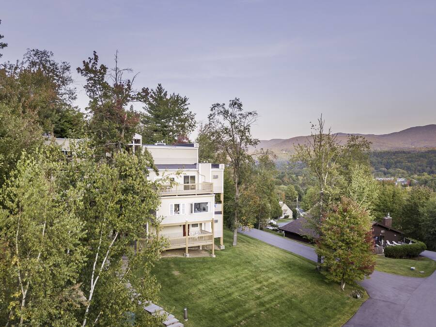 Un edificio de varios pisos con techo plano, rodeado de árboles y un paisaje montañoso.
