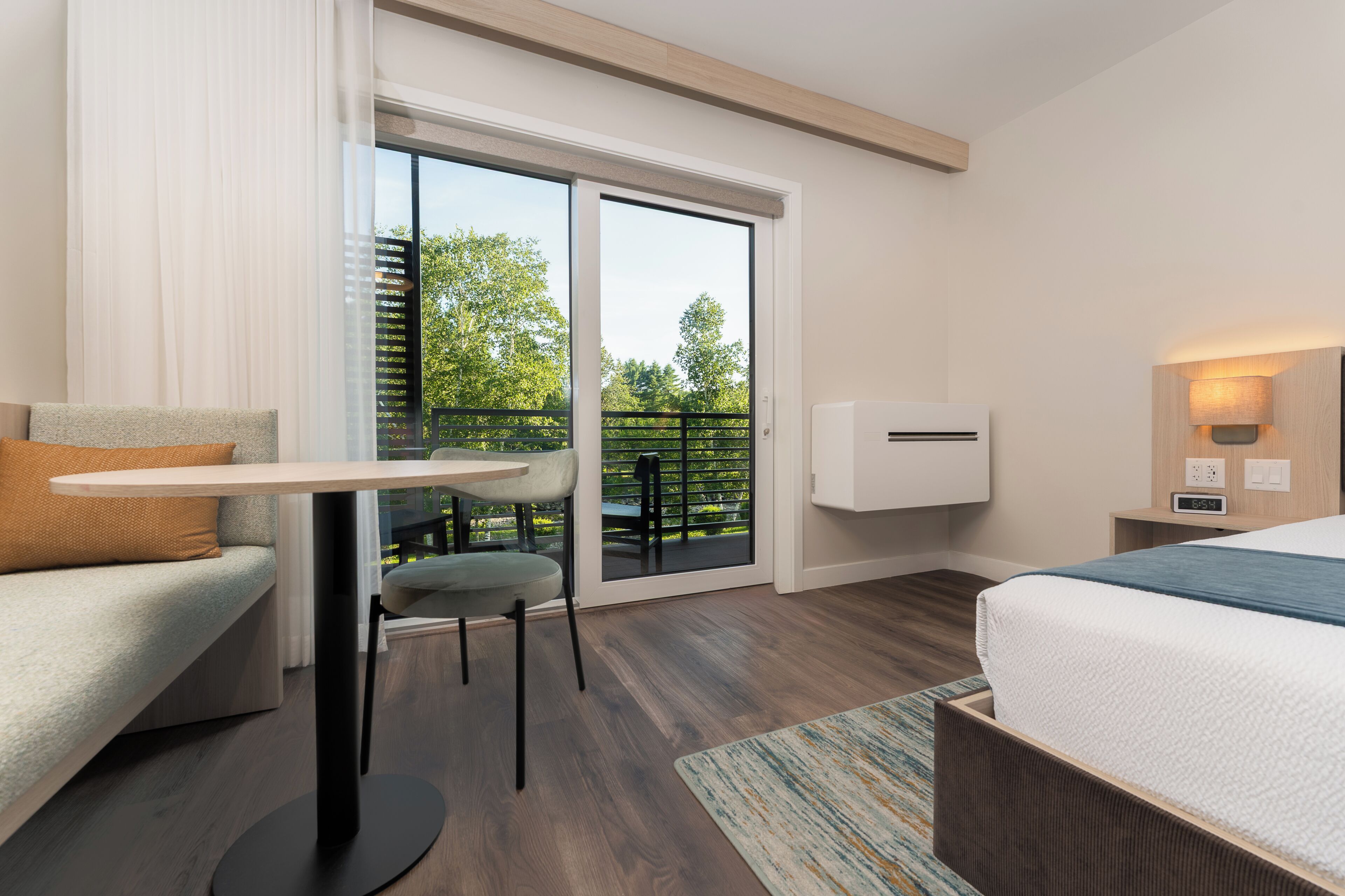 Habitación de hotel con cama, una mesita, una silla, un sofá, un balcón con vistas a árboles y un equipo de aire acondicionado instalado en la pared.