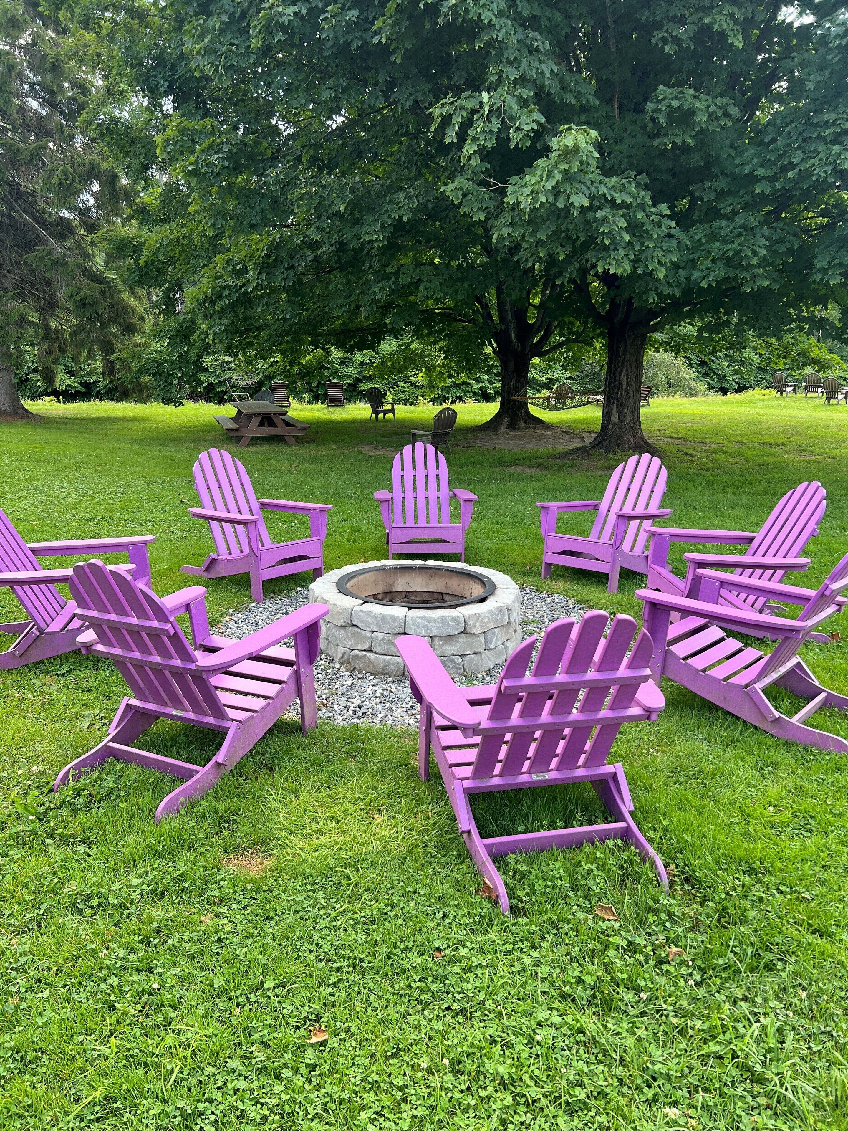 A circular seating arrangement with a stone fire pit in the center, surrounded by six purple Adirondack chairs, set on a grassy lawn with trees in the background.