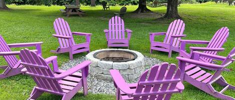 A circular seating arrangement with a stone fire pit in the center, surrounded by six purple Adirondack chairs, set on a grassy lawn with trees in the background.