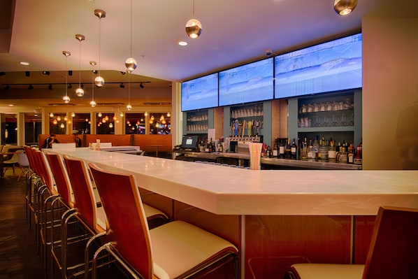A bar area with a long counter, multiple hanging lights, and a large screen displaying a cityscape. - Sun & Ski Inn And Suites (Stowe)