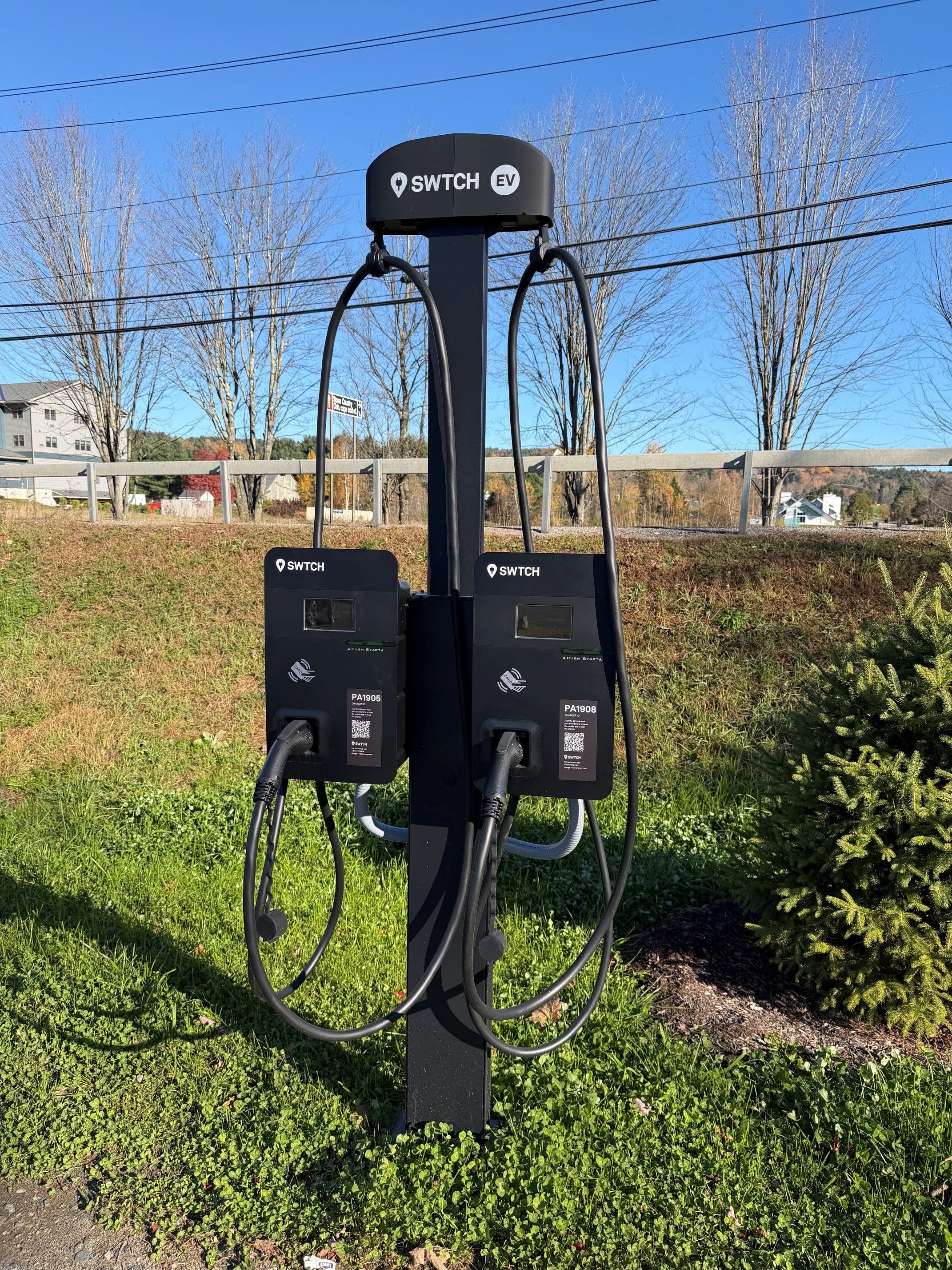 Una estación de carga para vehículos eléctricos con dos enchufes, identificada como SWITCH EV, en un área con césped, árboles y una valla al fondo.