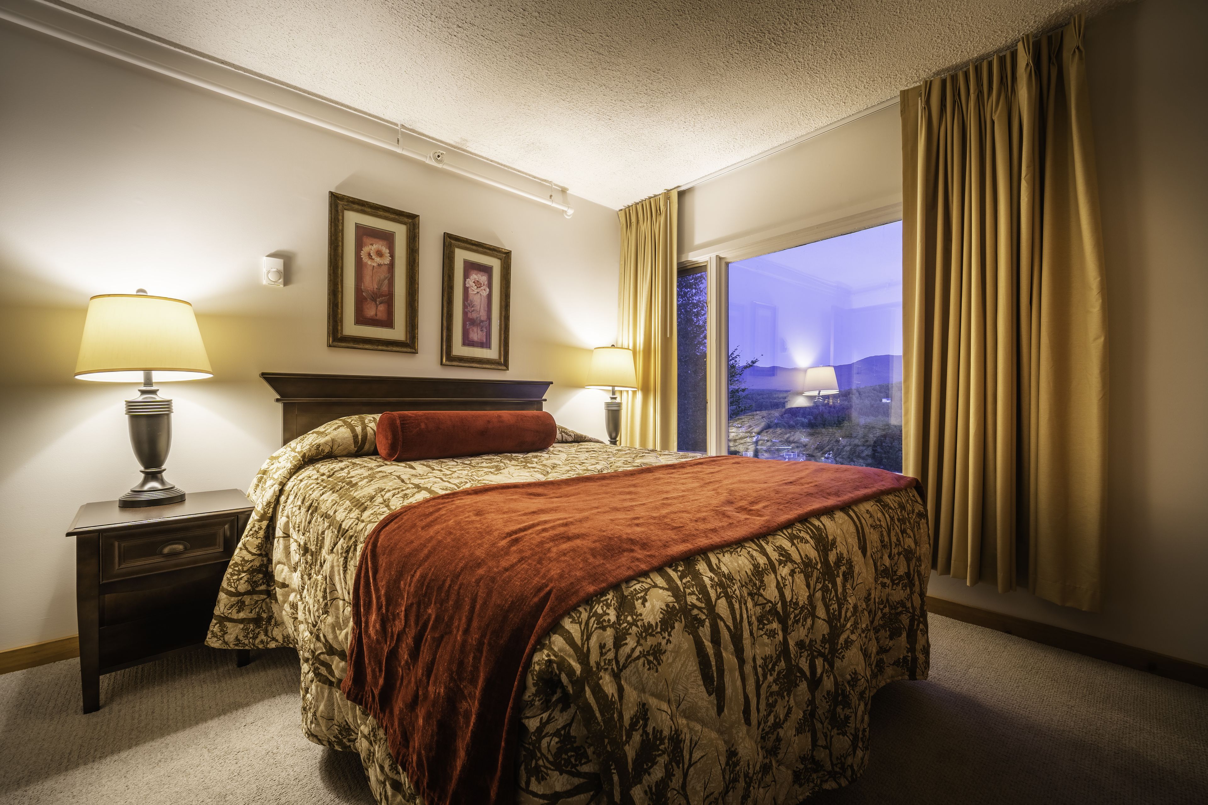 A hotel room with a bed, two bedside lamps, a nightstand, and a window with curtains.
