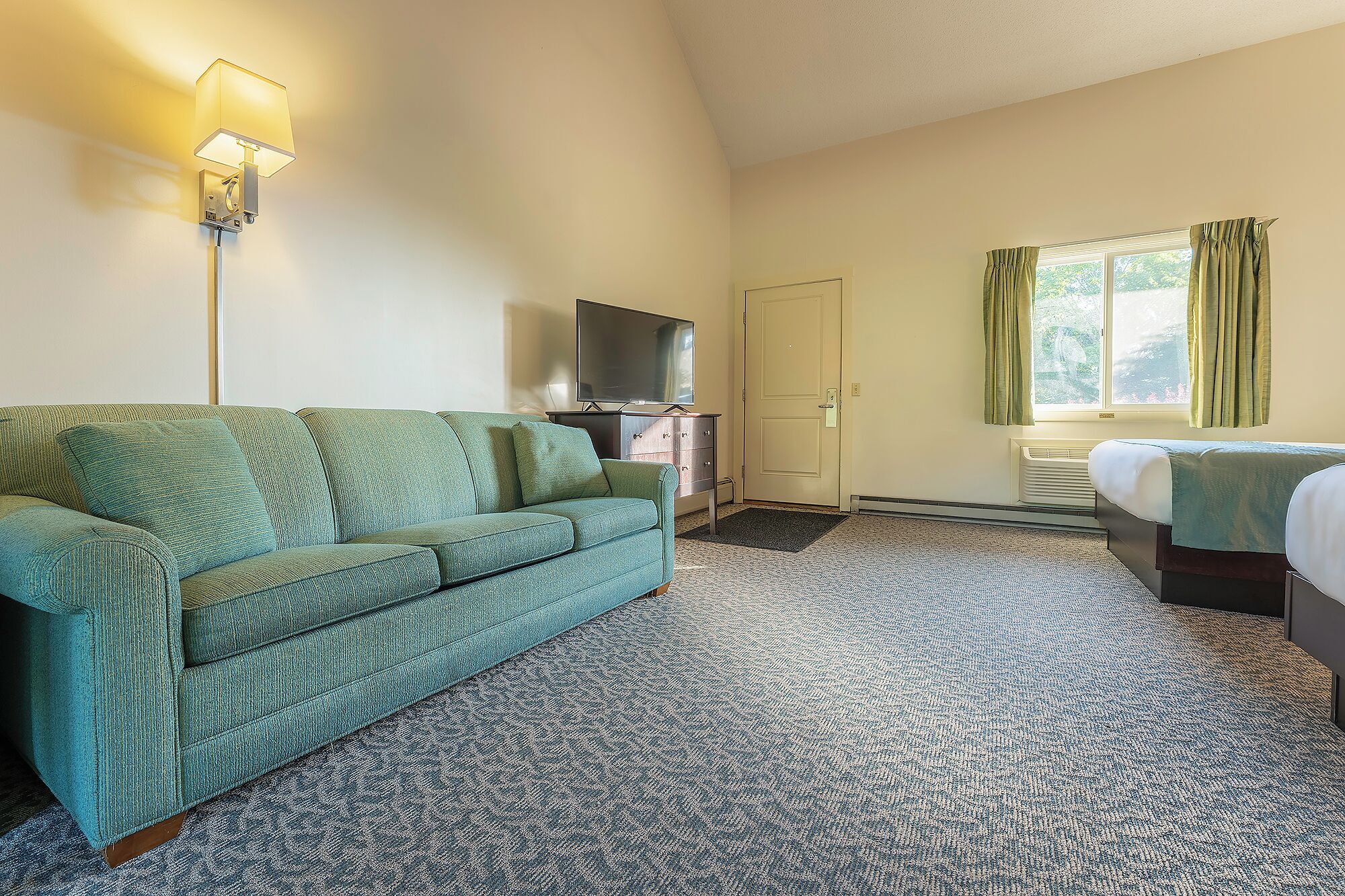 A hotel room with a green sofa, a TV, a bed, and a window with curtains.