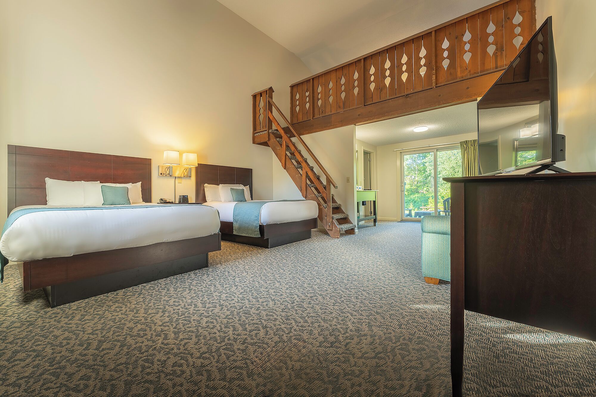 A hotel room with two beds, a wooden staircase, a desk, and a large window.