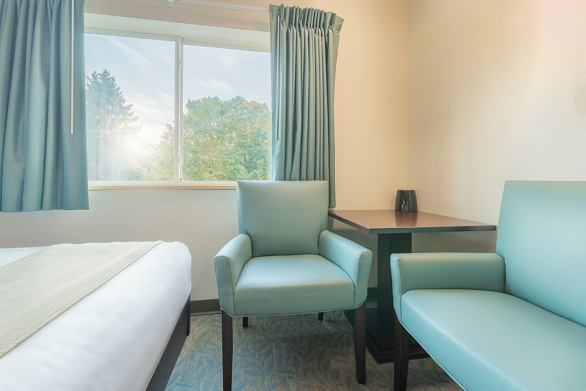 Habitación de hotel con una cama, dos sillones, una mesita y un ventanal con cortinas.