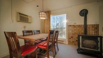 Une pièce chaleureuse avec une table à manger en bois, des chaises à coussins rouges, un luminaire suspendu, un poêle à bois et une fenêtre donnant sur les arbres.