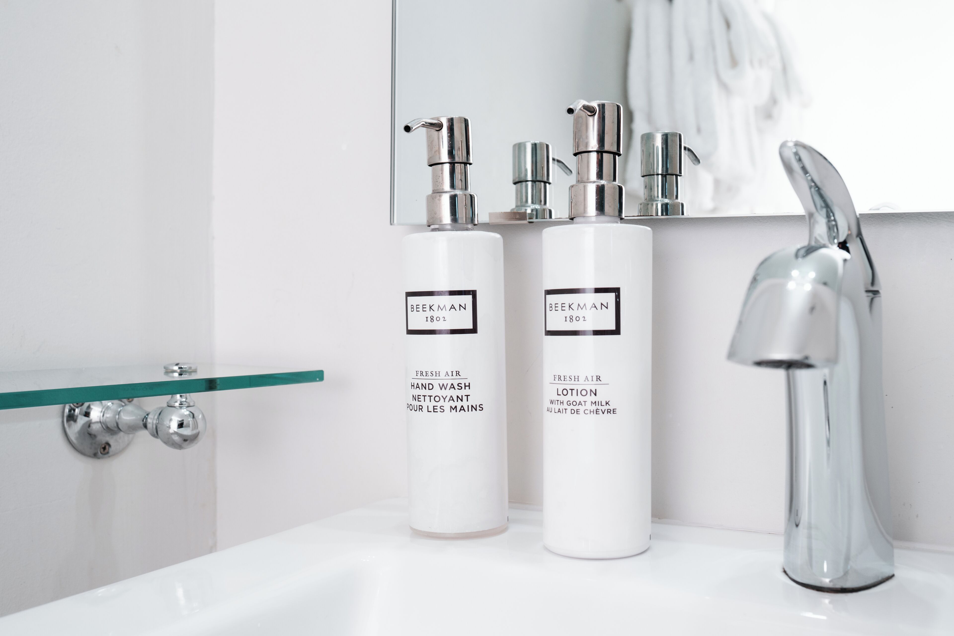 A bathroom with a white sink, two bottles of Beekman products, a glass shelf, and a chrome faucet.