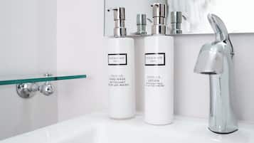 A bathroom with a white sink, two bottles of Beekman products, a glass shelf, and a chrome faucet.