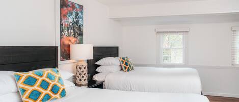 A hotel room with two beds, white bedding, a nightstand with a lamp, a framed artwork on the wall, and a window with blinds.