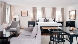 Una habitación de hotel con una cama, un sofá, una mesa de comedor y una silla. Hay dos mesitas de noche con lámparas y un cuadro en la pared.