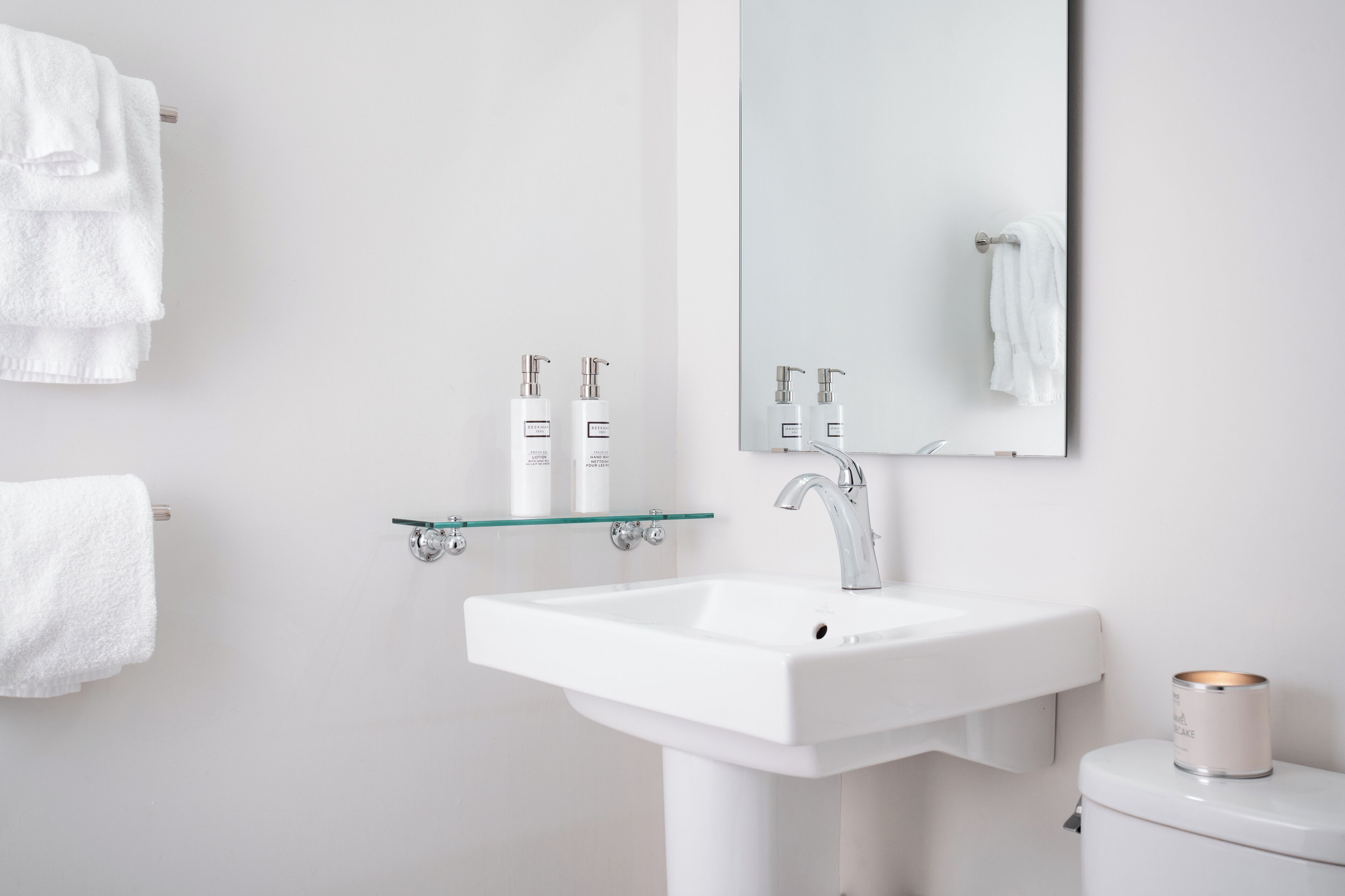 A modern bathroom with a white sink, a glass shelf with toiletries, and a toilet with a candle.
