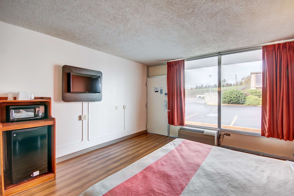 A hotel room with a bed, a television mounted on the wall, a small refrigerator, and a window with curtains.