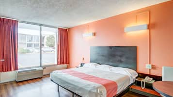 A hotel room with a large bed, a bedside table with a phone, a window with curtains, and a wall-mounted light fixture.