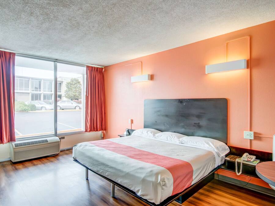A hotel room with a large bed, a bedside table with a phone, a window with curtains, and a wall-mounted light fixture.
