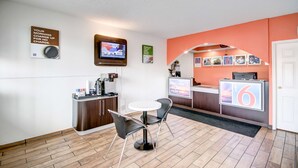 Zona de recepción de un hotel, con mostrador, pantalla de televisión, cafetera y un área de descanso con dos sillas.