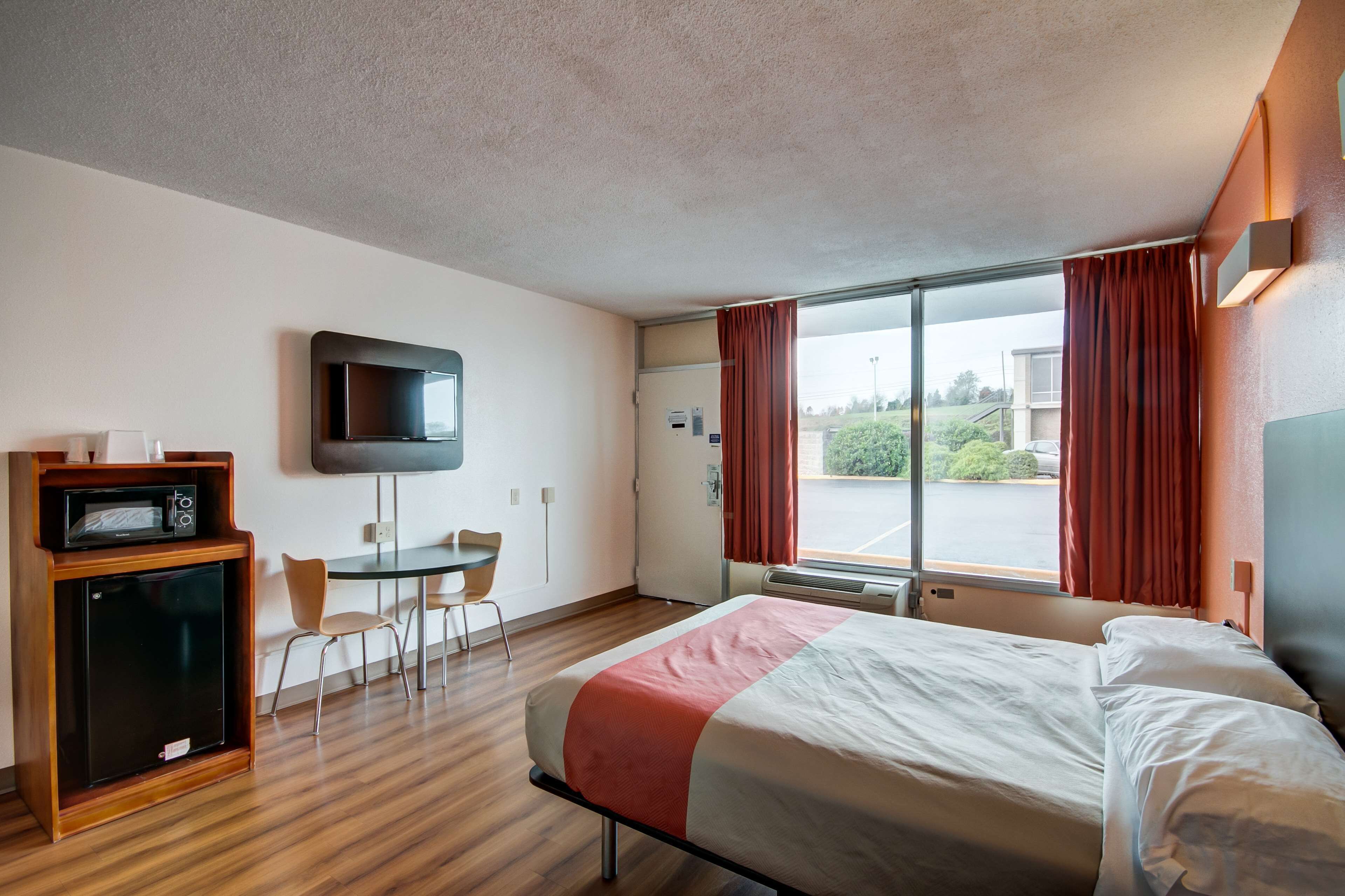 Et hotelværelse med en seng, et fjernsyn ophængt på væggen, et skrivebord med stol og et vindue med gardiner.