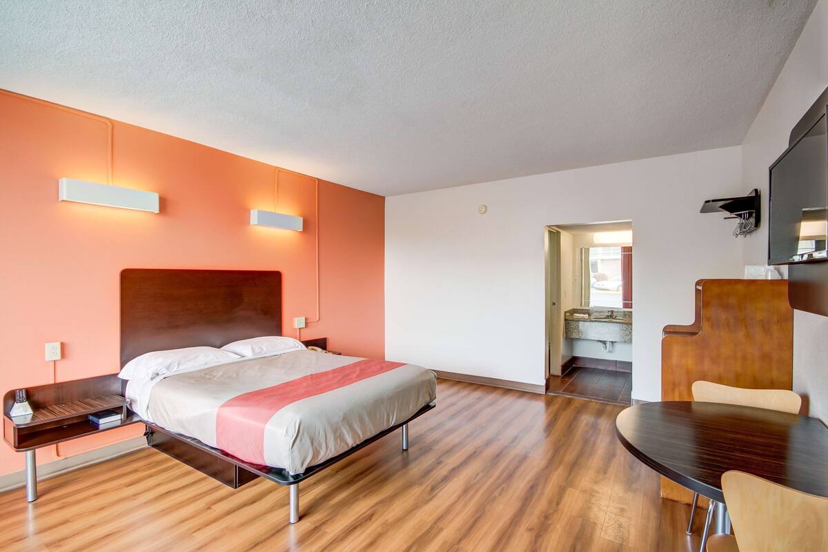A modern hotel room with a wooden floor, a bed with a red and white bedspread, a bedside table with a remote control, and a wooden chair at a small table.