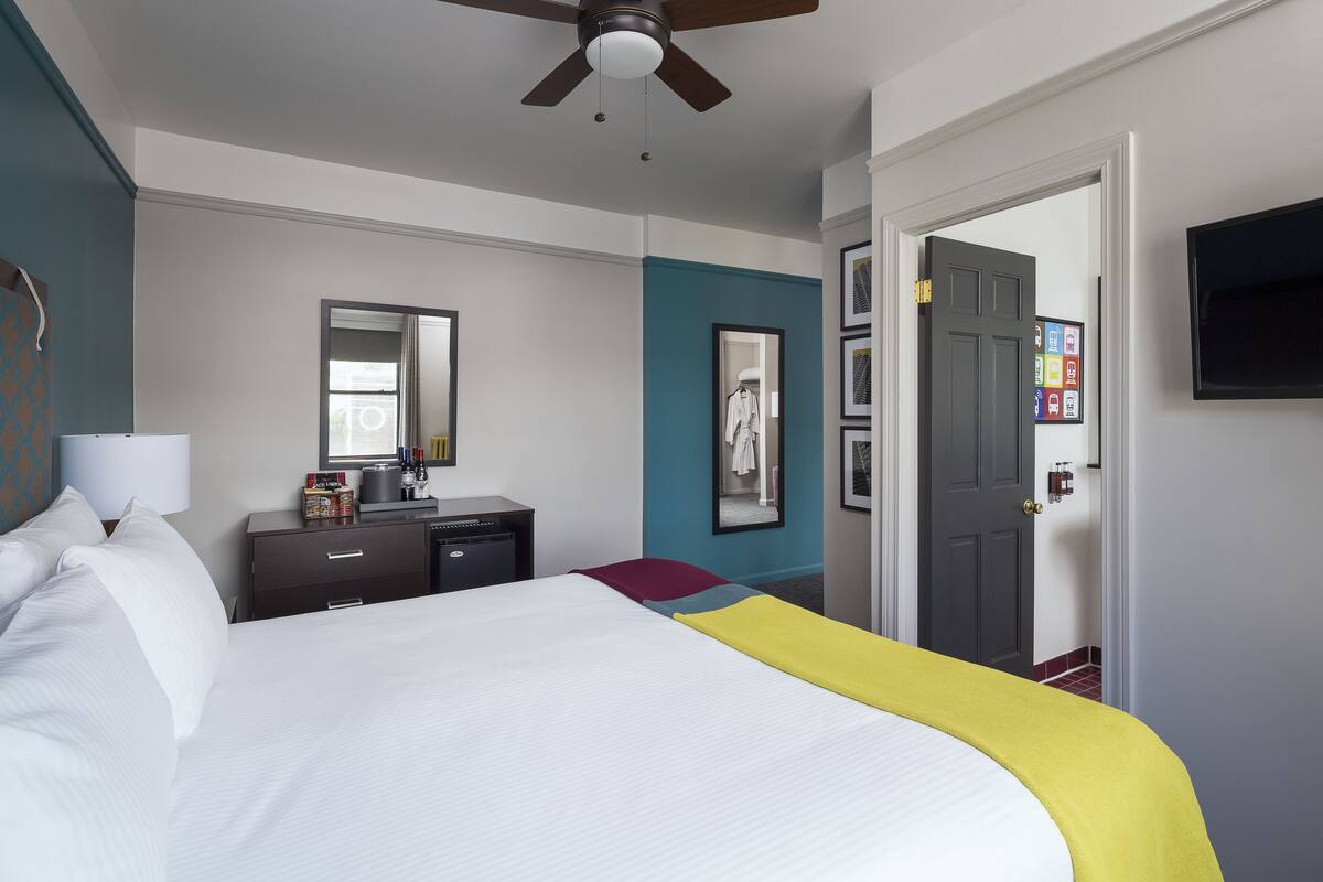 A hotel room with a bed, a desk, a chair, a wardrobe, a mirror, a television, and a door with a lock.
