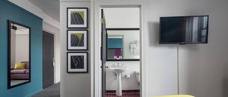A hotel room with a bed, a television, a sink, and framed artwork on the wall.