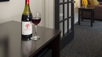 Habitación de hotel con un escritorio de madera oscura, una botella de vino y una copa sobre la mesa. Se ve una puerta con paneles de vidrio que da a otra habitación.