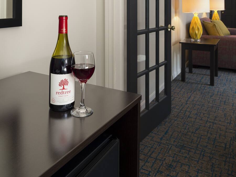 Habitación de hotel con un escritorio de madera oscura, una botella de vino y una copa sobre la mesa. Se ve una puerta con paneles de vidrio que da a otra habitación.