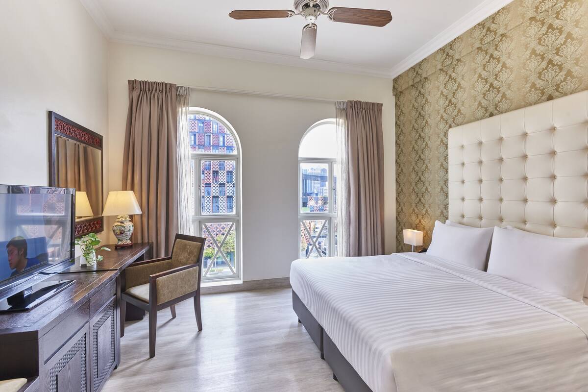 A hotel room with a bed, a desk with a television, two windows with curtains, a ceiling fan, and a decorative wall.