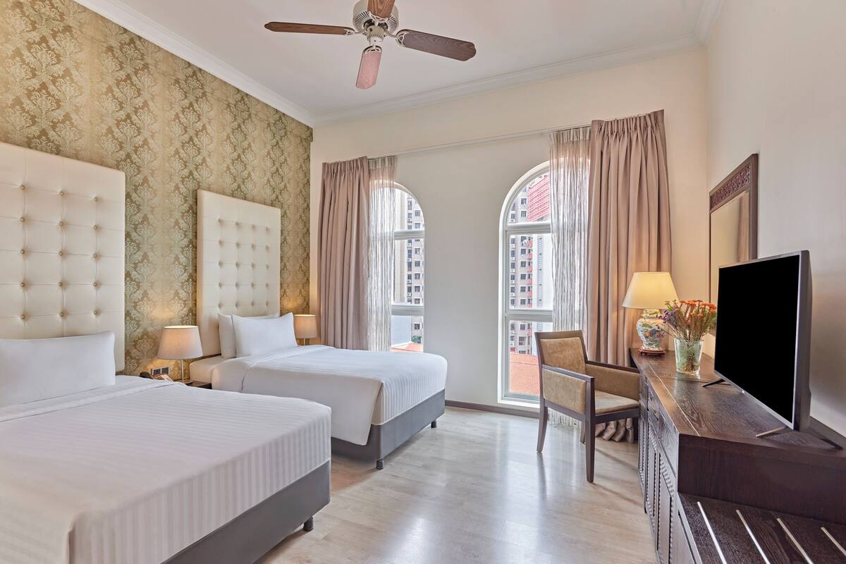 A hotel room with two beds, a ceiling fan, a TV, and a view of the city through large windows.