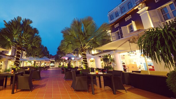 An outdoor dining area with tables and chairs, palm trees, and buildings with illuminated signs. - Village Hotel Albert Court by Far East Hospitality (Singapore)