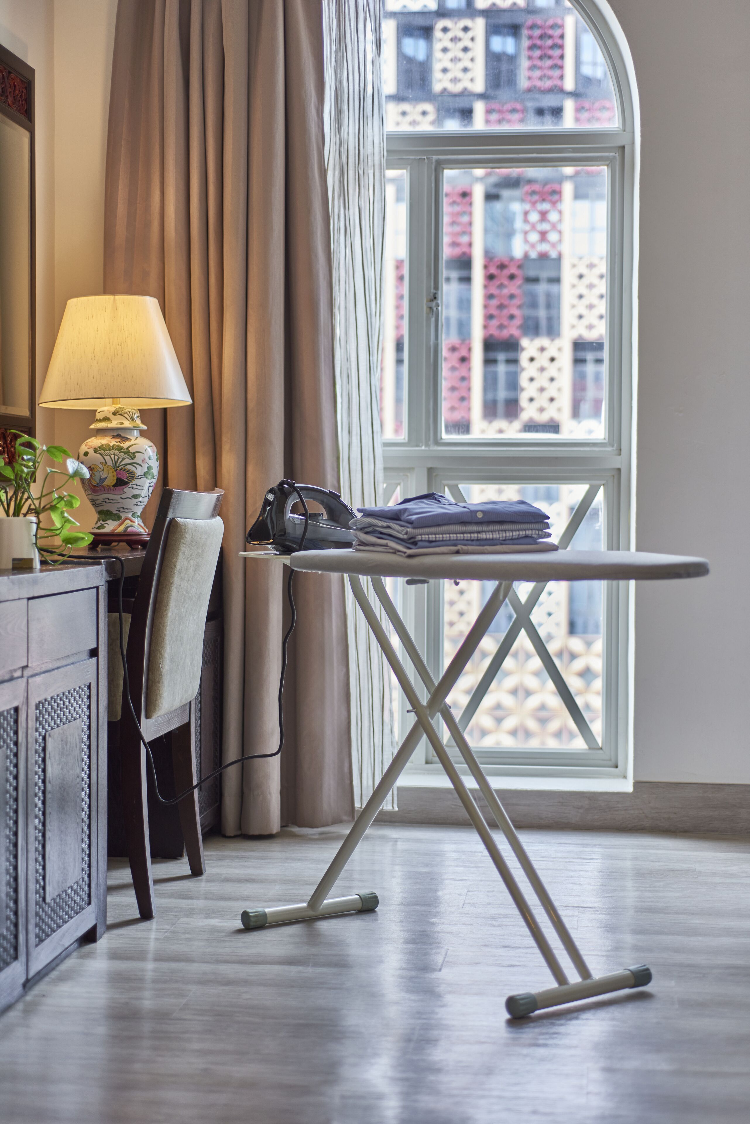 A neatly arranged workspace with an ironing board, a lamp, and a chair near a window with a view of patterned buildings.
