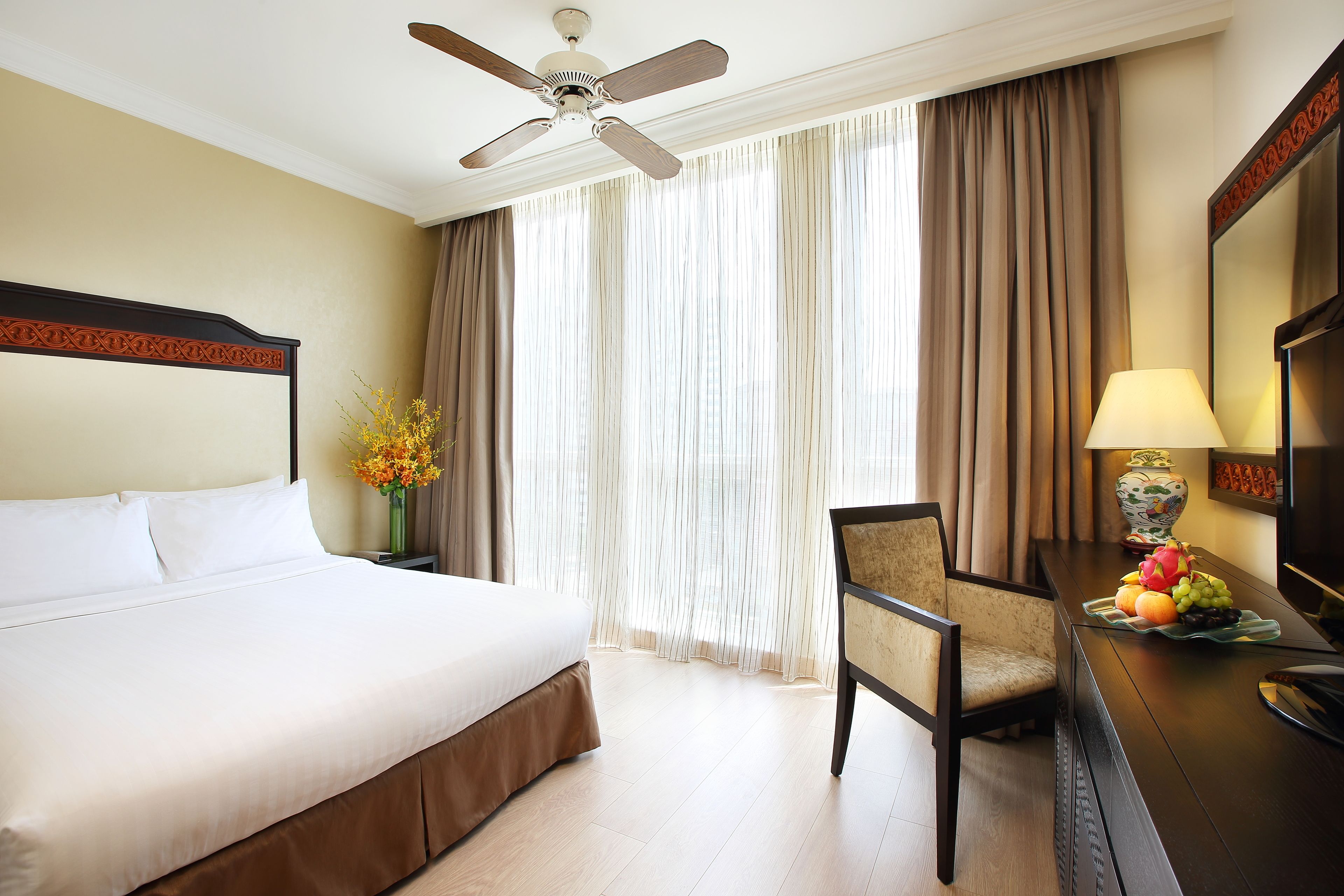 Habitación de hotel con una cama, un escritorio con lámpara, una silla, un frutero y un ventilador de techo.