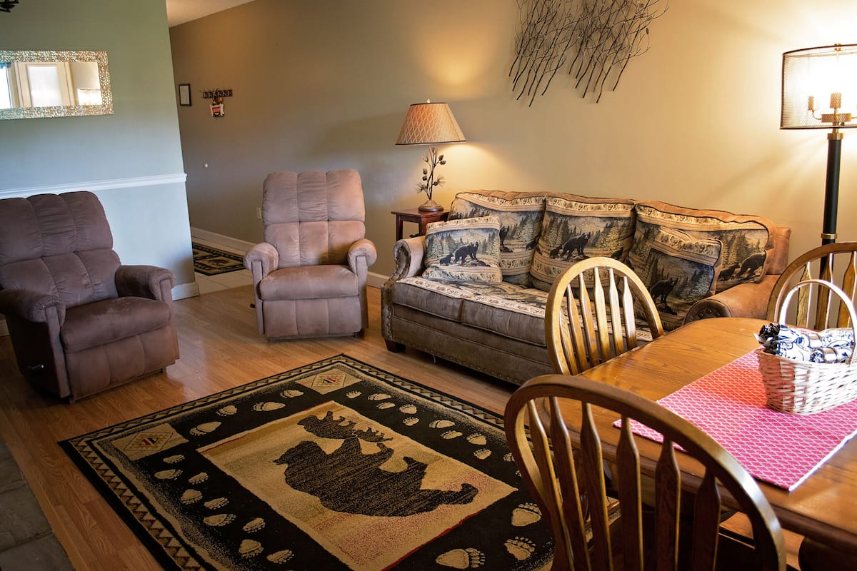 A living room with a sofa, two armchairs, a dining table with chairs, a patterned rug, and a lamp.
