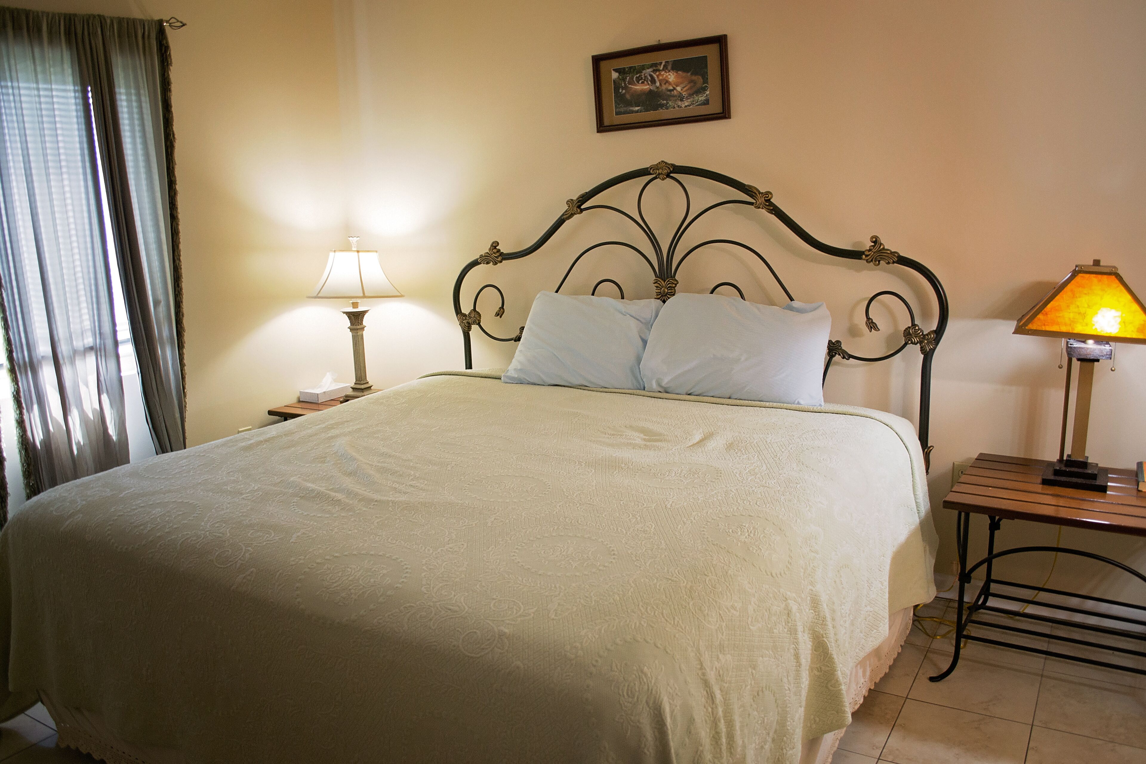 A neatly made bed with a headboard, two bedside tables with lamps, a framed picture on the wall, and a window with curtains.