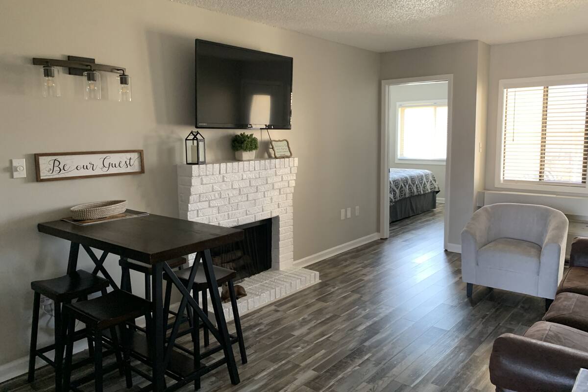 A modern living room with a fireplace, a dining area with bar stools, a flat-screen TV mounted above the fireplace, and a cozy seating area with a chair and a sofa.