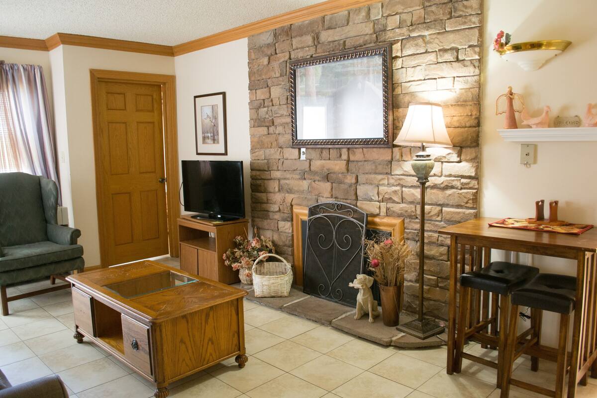 A cozy living room with a stone fireplace, a television, a wooden coffee table, and a dining area with bar stools.
