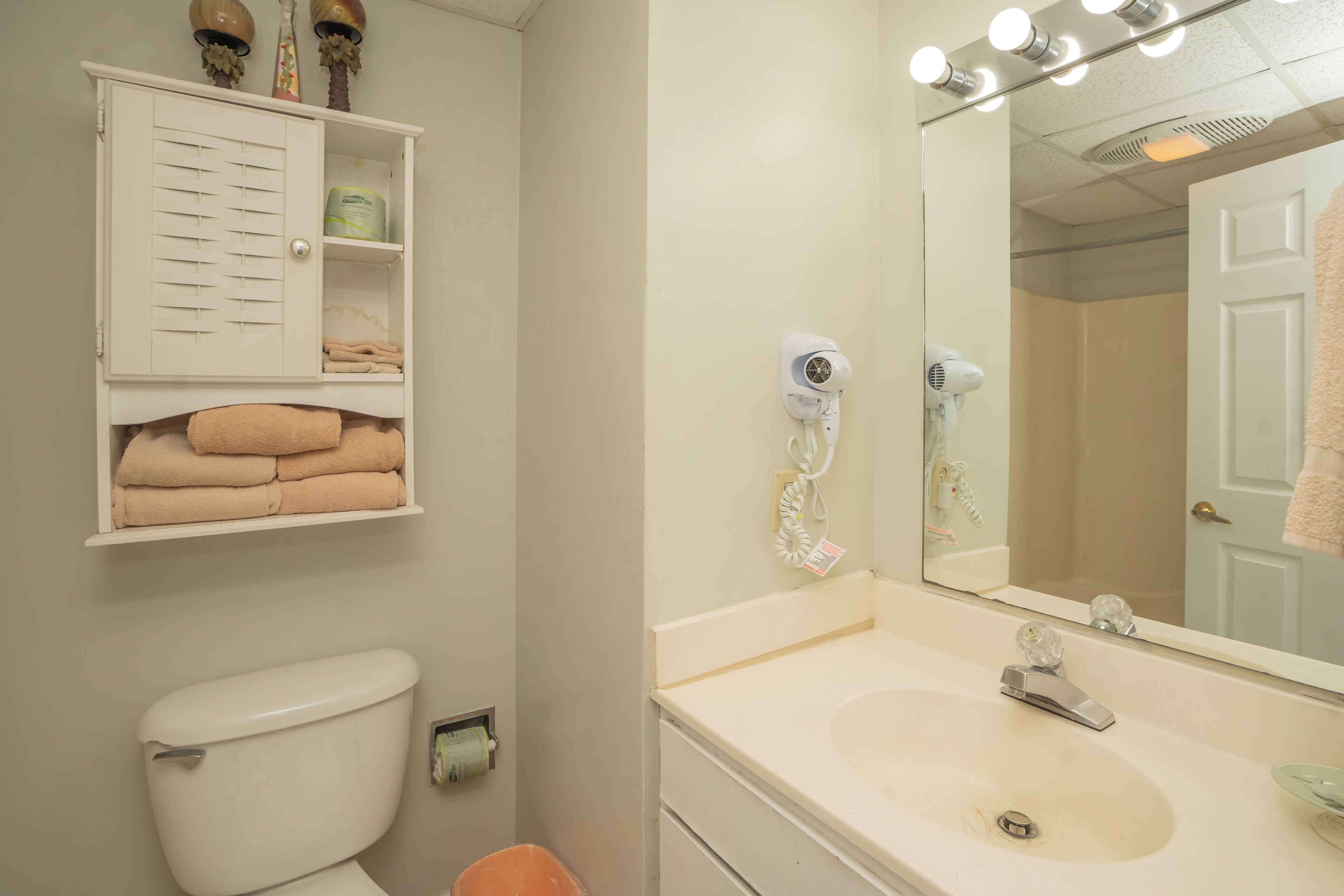 A bathroom with a toilet, a sink, a mirror with lights, a towel rack, and a cabinet with towels.