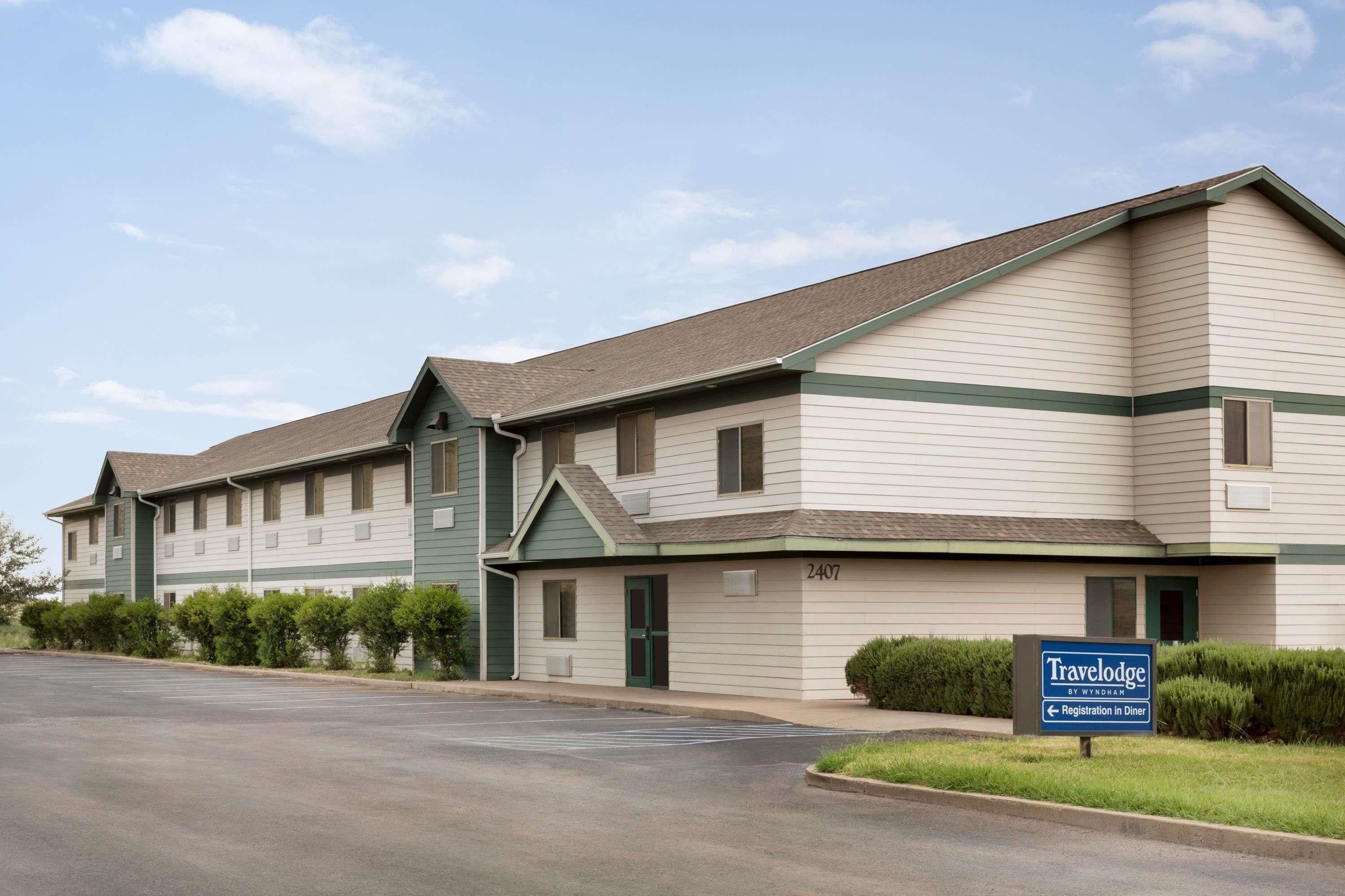 A Travelodge motel with a green and white exterior, a parking lot, and a sign indicating a registration area for the dining room.