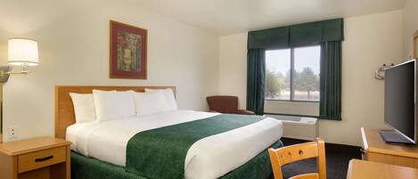 A hotel room with a large bed, a desk with a computer, a chair, a lamp, and a window with curtains.