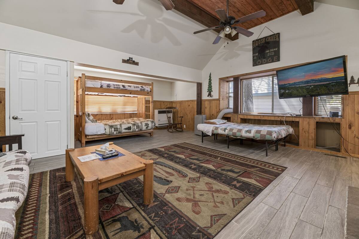 Ein gemütliches Chalet-Interieur mit Holzbalkendecke, einem großen Fernseher, einer hölzernen Sitzbank und einem gemusterten Teppich.