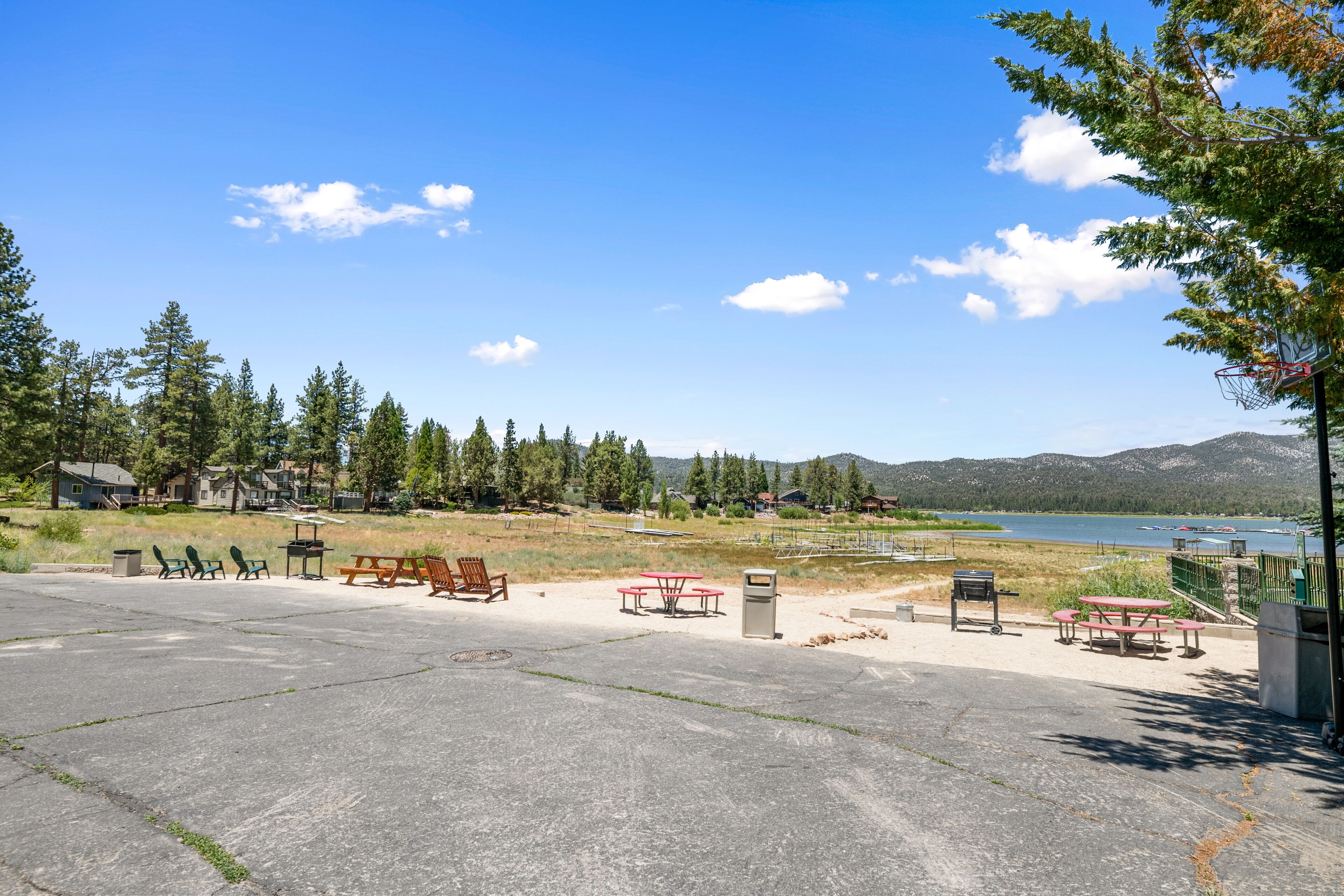 Un espacio al aire libre con mesas de picnic y bancos, un aro de baloncesto y vistas a un lago y montañas.