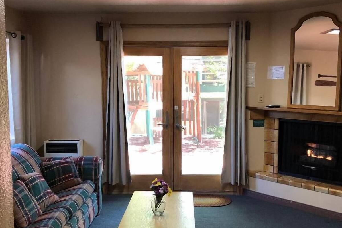 Ein Wohnzimmer mit einer Couch, einem Couchtisch mit einer Vase, einem Kamin und einer Schiebeglastür, die zu einem Außenbereich führt.