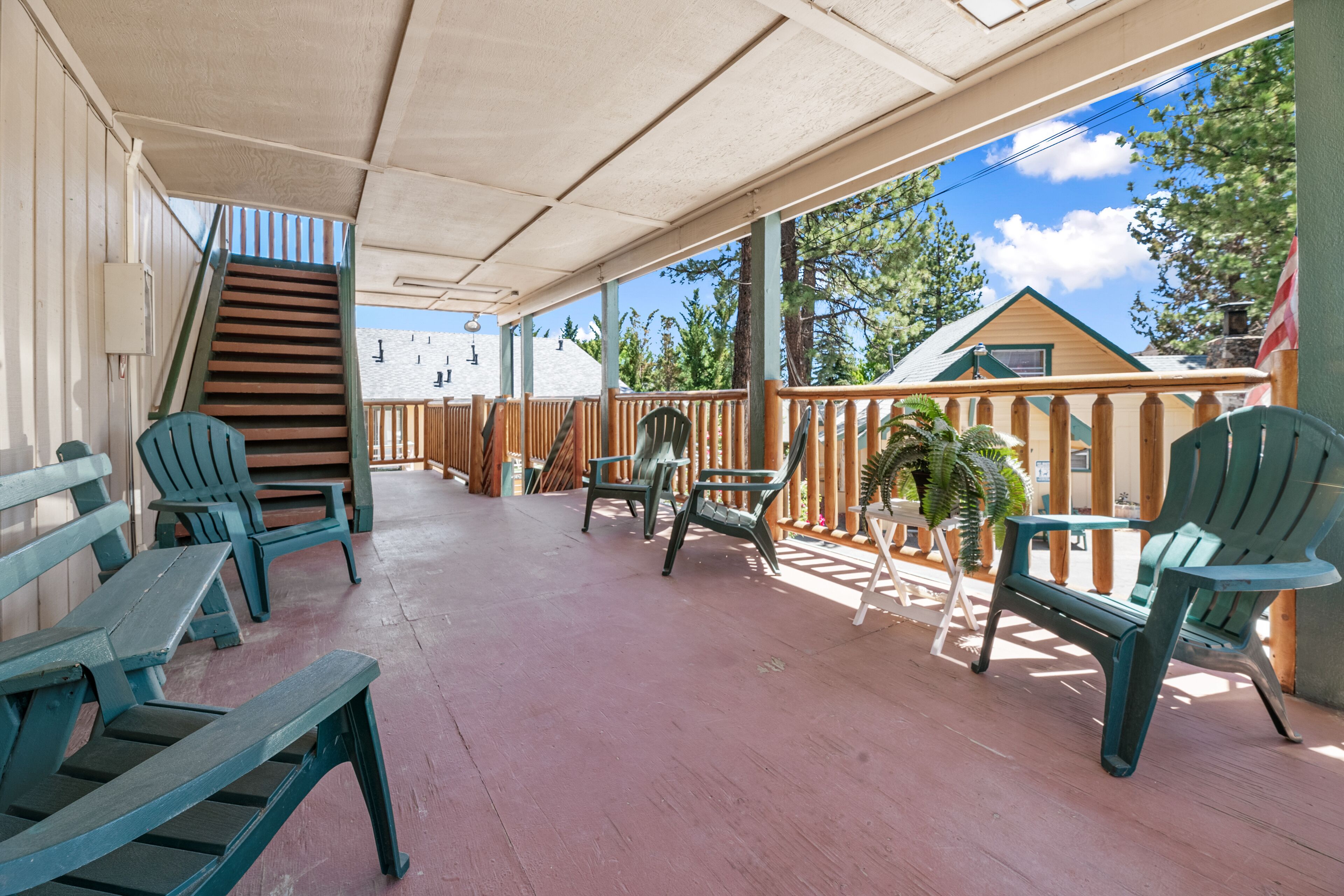 A covered outdoor area with wooden benches and chairs, a staircase leading to an upper level, and a view of a house and trees in the background.