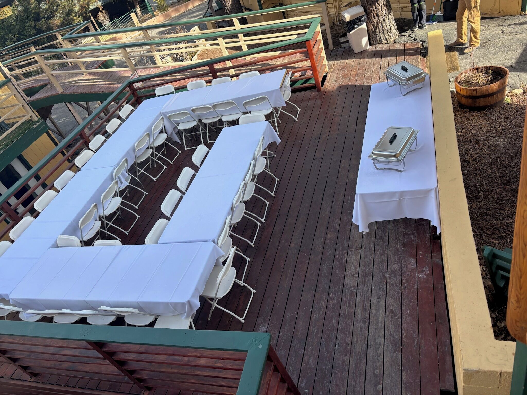 A covered outdoor area with long white tables and chairs, wooden deck flooring, and a metal railing.
