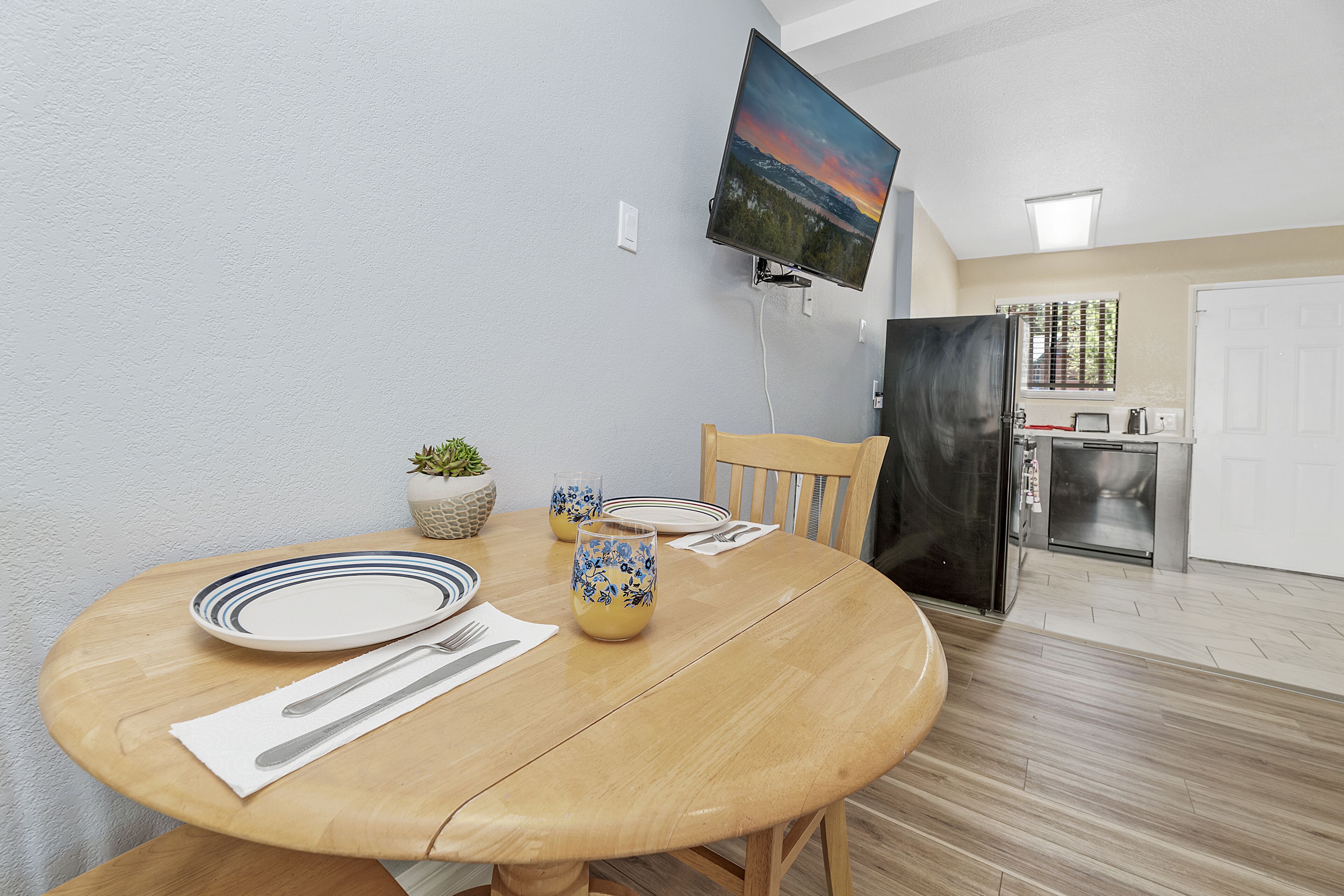Un comedor con una mesa de madera preparada para dos comensales, un televisor de pantalla plana montado en la pared y una cocina al fondo.
